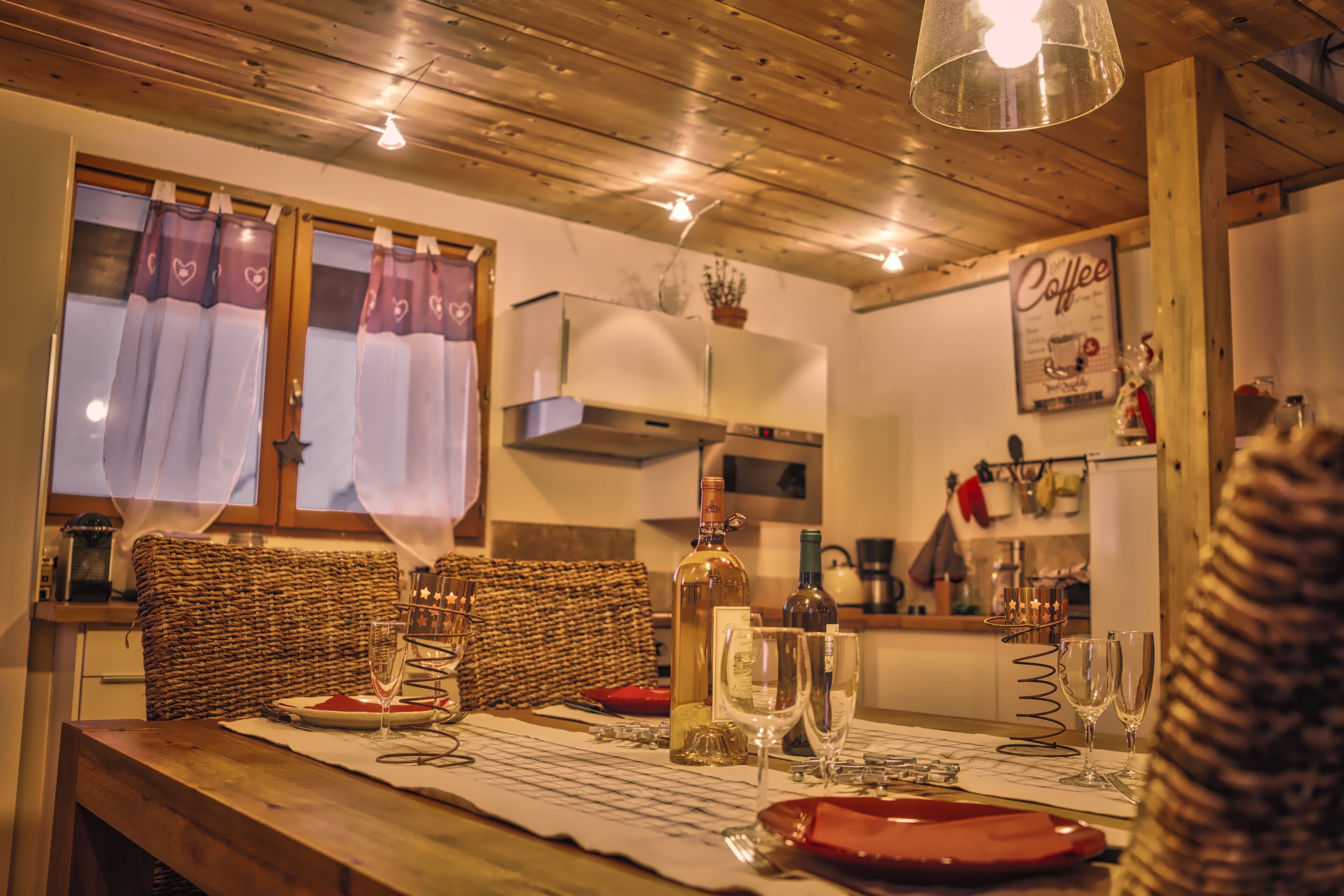 A cozy dining area with a wooden table set for two, a wicker chair, and a kitchen in the background.