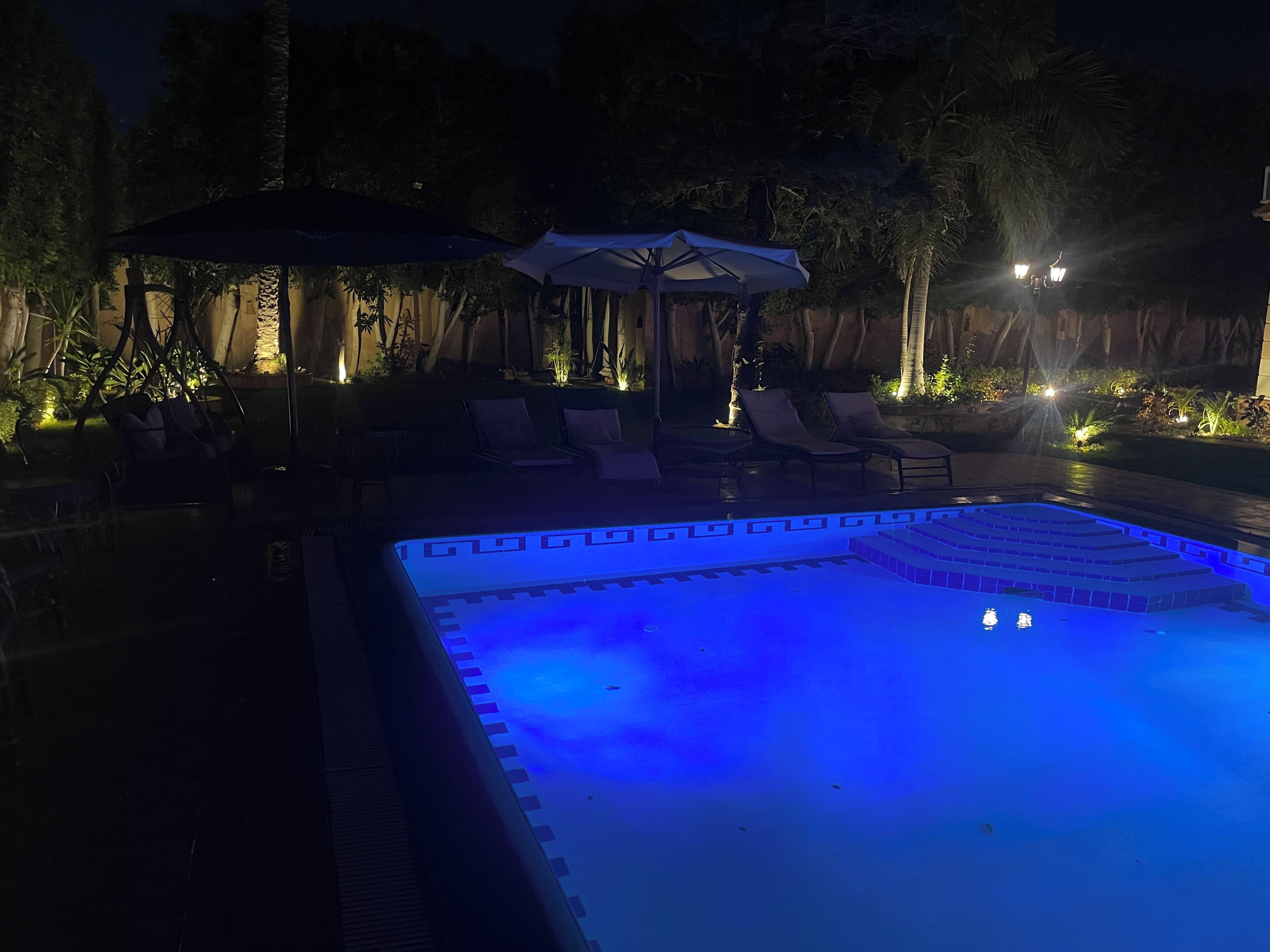 A nighttime swimming pool area with lounge chairs and umbrellas, surrounded by trees and illuminated by ambient lighting.