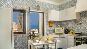 A compact kitchen with a view of the sea, featuring a white table with stools, a microwave, a stove, and a clock on the wall.