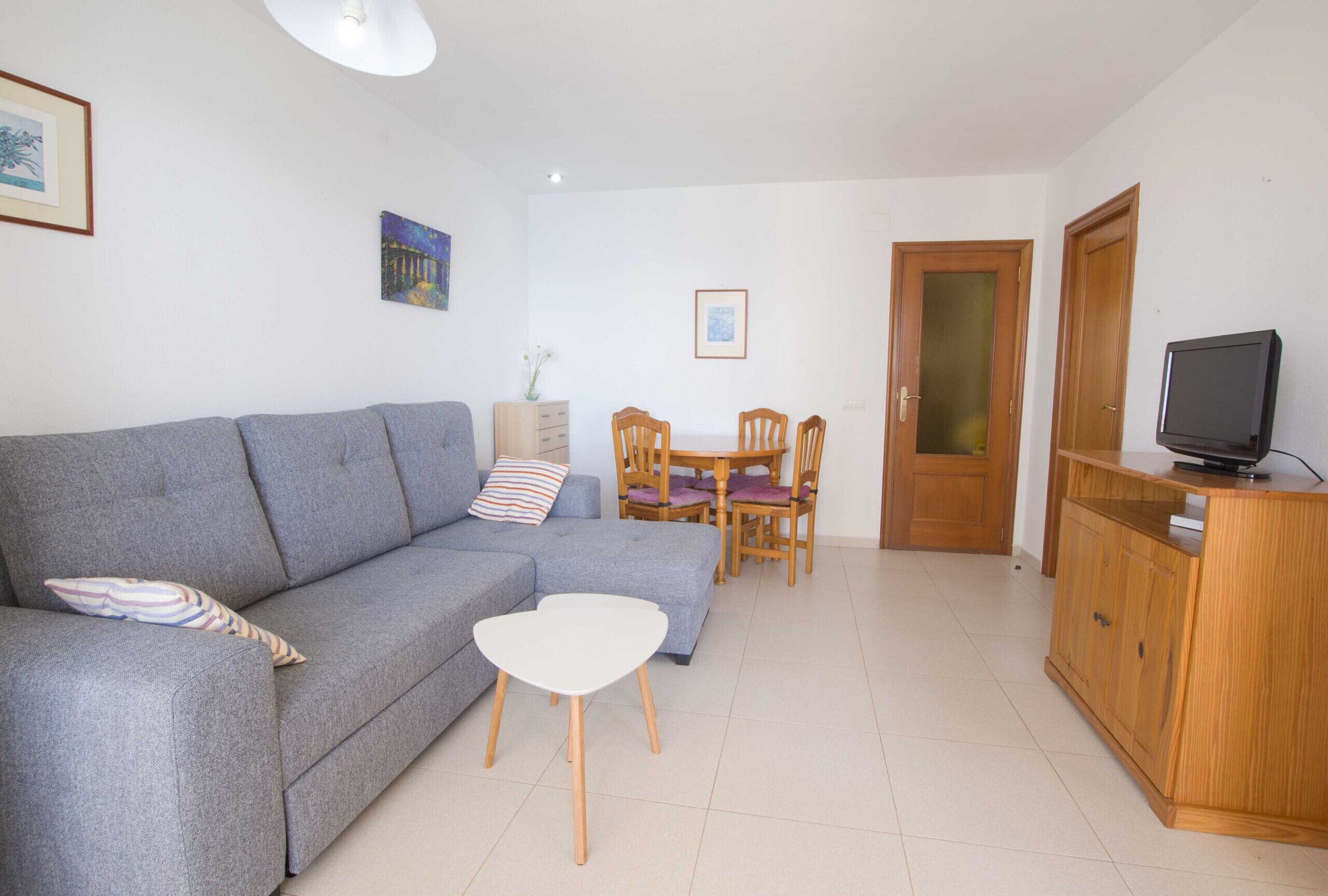 A living room with a grey sofa, a small white table, wooden chairs, a wooden cabinet, and a television.