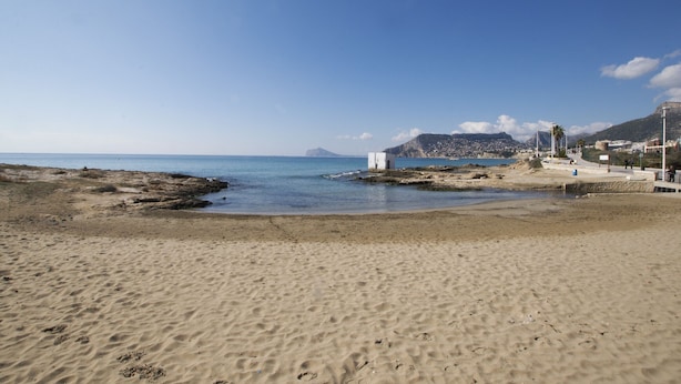 Une plage de sable avec un promontoire rocheux, un bâtiment blanc et une montagne en arrière-plan.