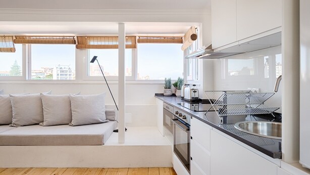 A modern kitchen with a built-in oven, a sink, and a countertop. There is a window with a view of the city, and a sofa with cushions in the adjacent living area. - Romantic Gem in Graça (Lisboa)