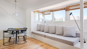 A modern living room with a built-in bench, a round dining table with chairs, and a large window with bamboo blinds. - Romantic Gem in Graça (Lisboa)