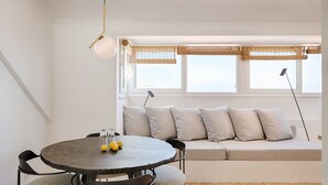 A modern living room with a round dining table, a sofa with cushions, and a large window with blinds. - Romantic Gem in Graça (Lisboa)