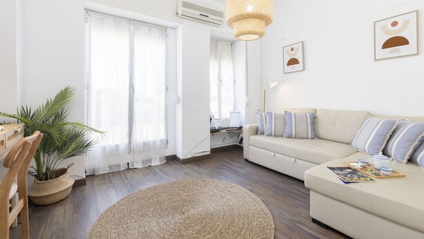 Ein modernes Wohnzimmer mit einem beigefarbenen Sofa, einem runden Teppich, einem Holzstuhl, einer Topfpflanze und einer Pendelleuchte.