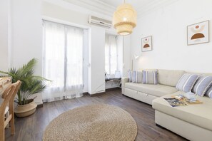 A modern living room with a beige sofa, a round woven rug, a wooden chair, a potted plant, and a hanging light fixture.