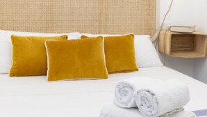 Ein Bett mit weißer Bettwäsche und zwei senfgelben Kissen, ein aufgerolltes weißes Handtuch sowie ein kleiner Holfnachtisch mit einem Buch.