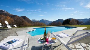 Un espace aménagé au bord de la piscine, avec des chaises longues, une table sur laquelle se trouvent une bouteille, des verres et un bol de collations, le tout dans un cadre dominé par des montagnes.