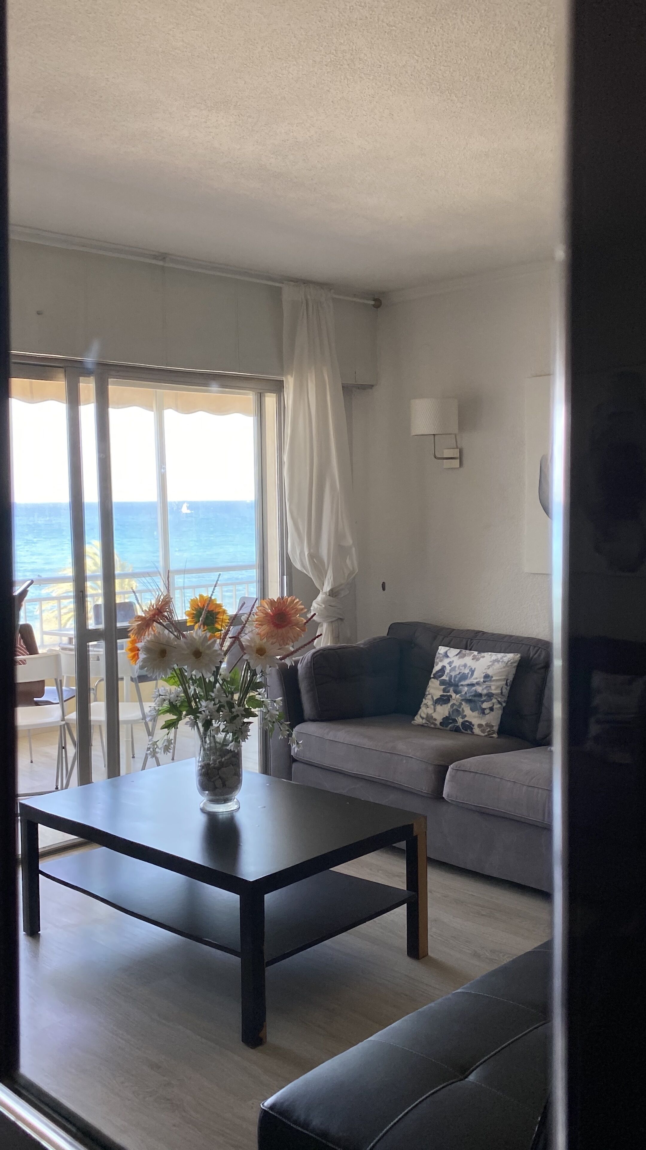 A living room with a sofa, a coffee table with a vase of flowers, a view of the ocean through large windows, and a curtain.
