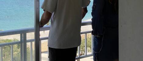A man and a woman standing by a window overlooking a coastal view with mountains in the background.