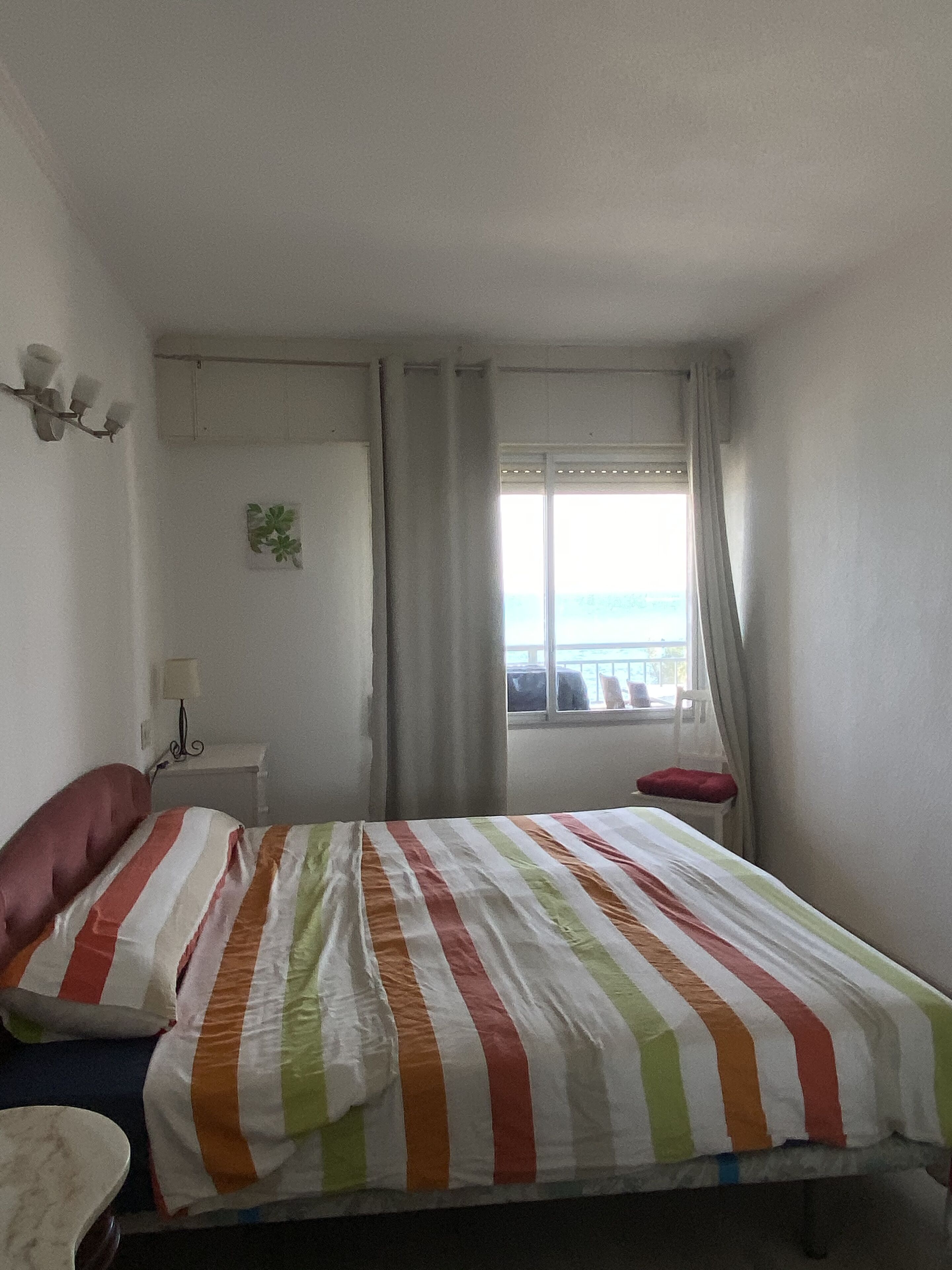 Een slaapkamer met een bed, nachtkastje, lamp, raam met gordijnen en uitzicht op zee.