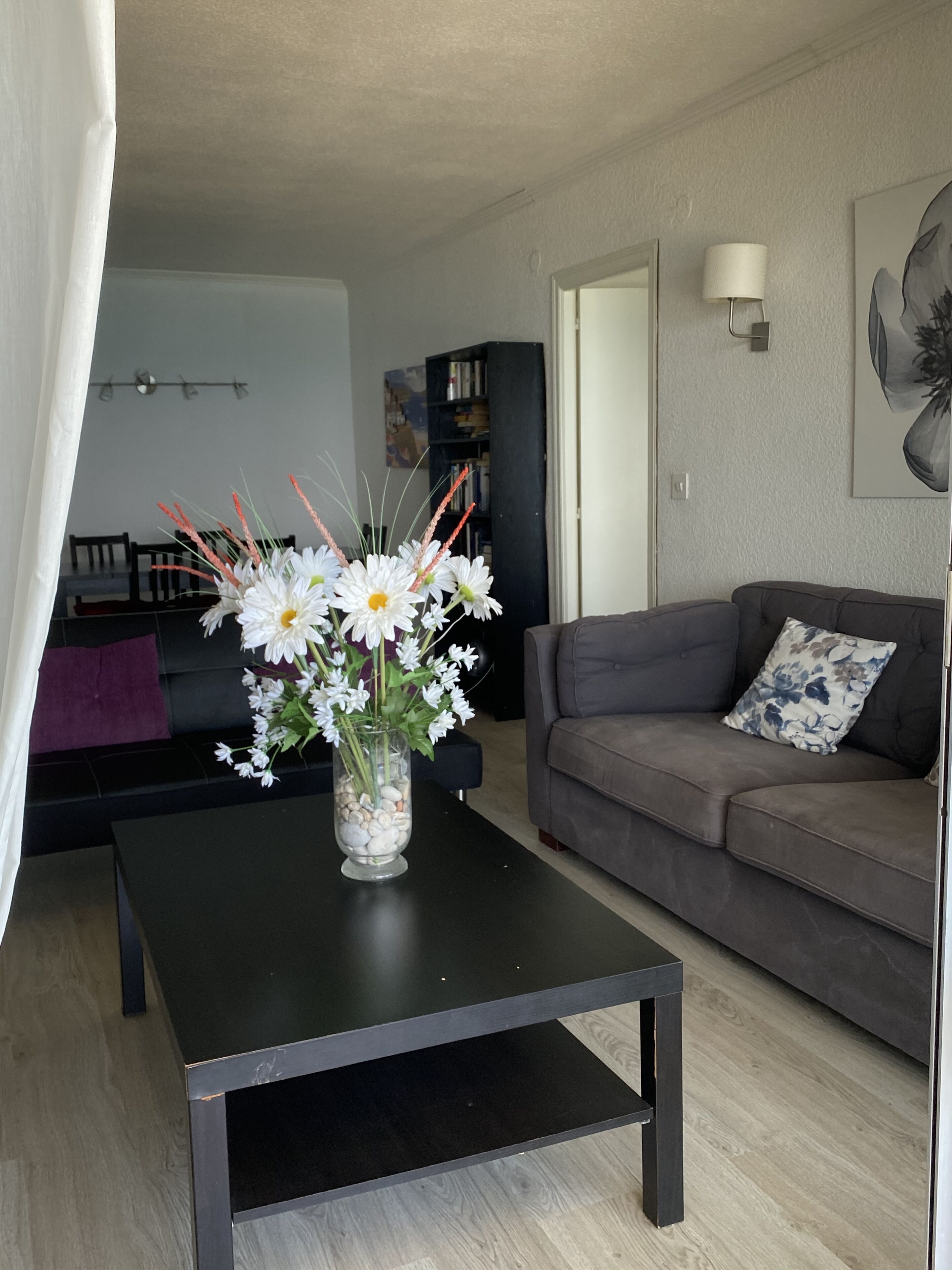 A living room with a dark coffee table, a grey sofa with a floral pillow, and a vase with white flowers on the table.