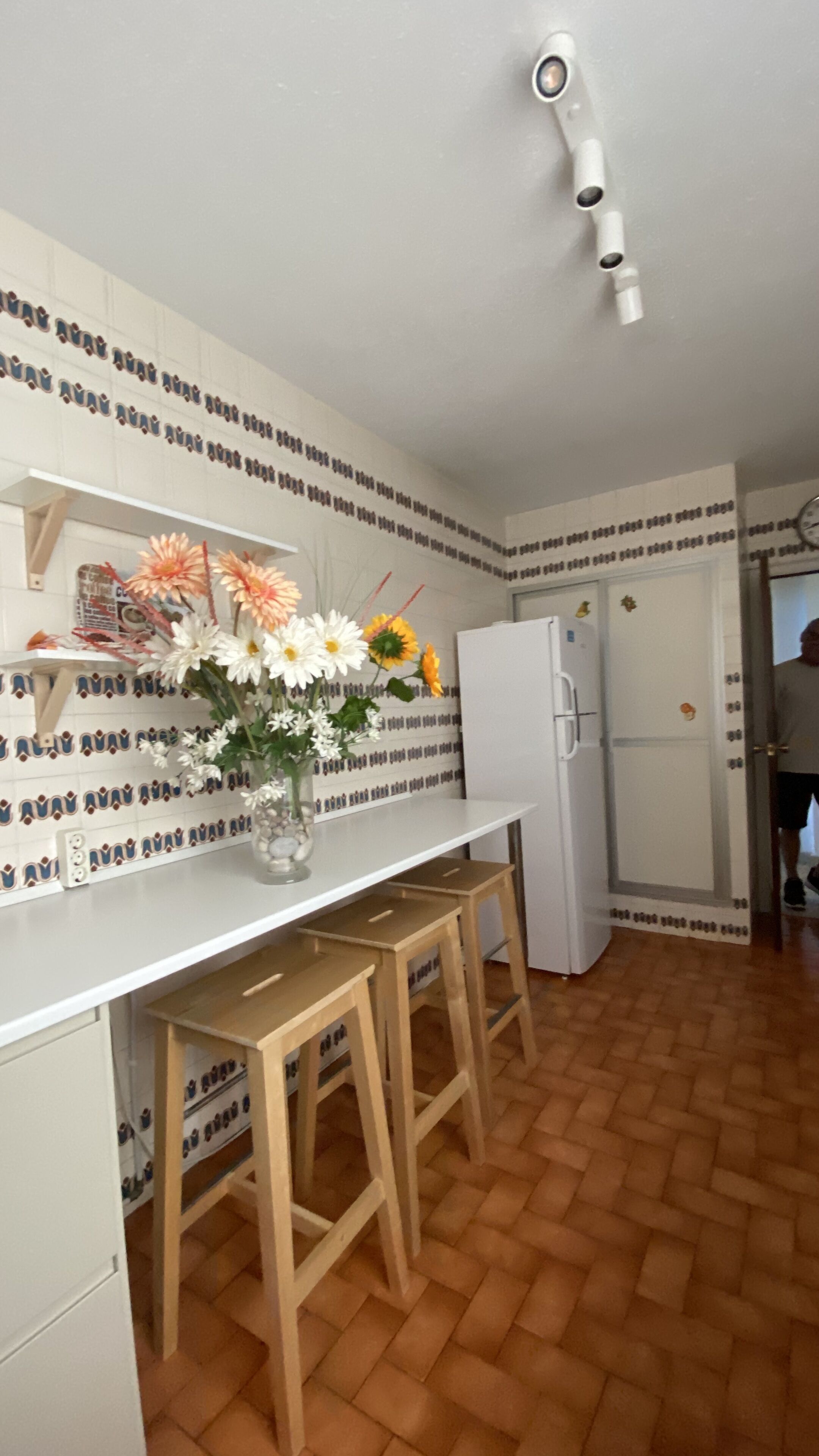 A kitchen with a white countertop, a wooden stool, a white refrigerator, and a wall with patterned tiles.