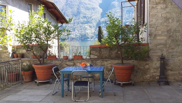 Un patio exterior con una mesa y sillas, plantas en macetas y vistas a un lago y montañas.
