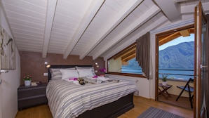 A bedroom with a large bed, a nightstand, a window with a view of mountains, and a balcony with a chair.