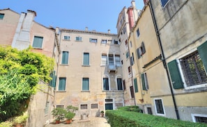 A narrow street with buildings on either side, featuring balconies and a small garden area.