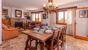 Comedor con una mesa de madera dispuesta para cuatro personas, rodeada de sillas. Hay una chimenea con manto de piedra, un sofá y un televisor al fondo.