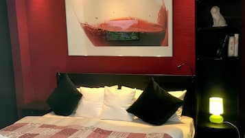 Quarto de hotel com cama, quadro, mesa de cabeceira e estante de livros.