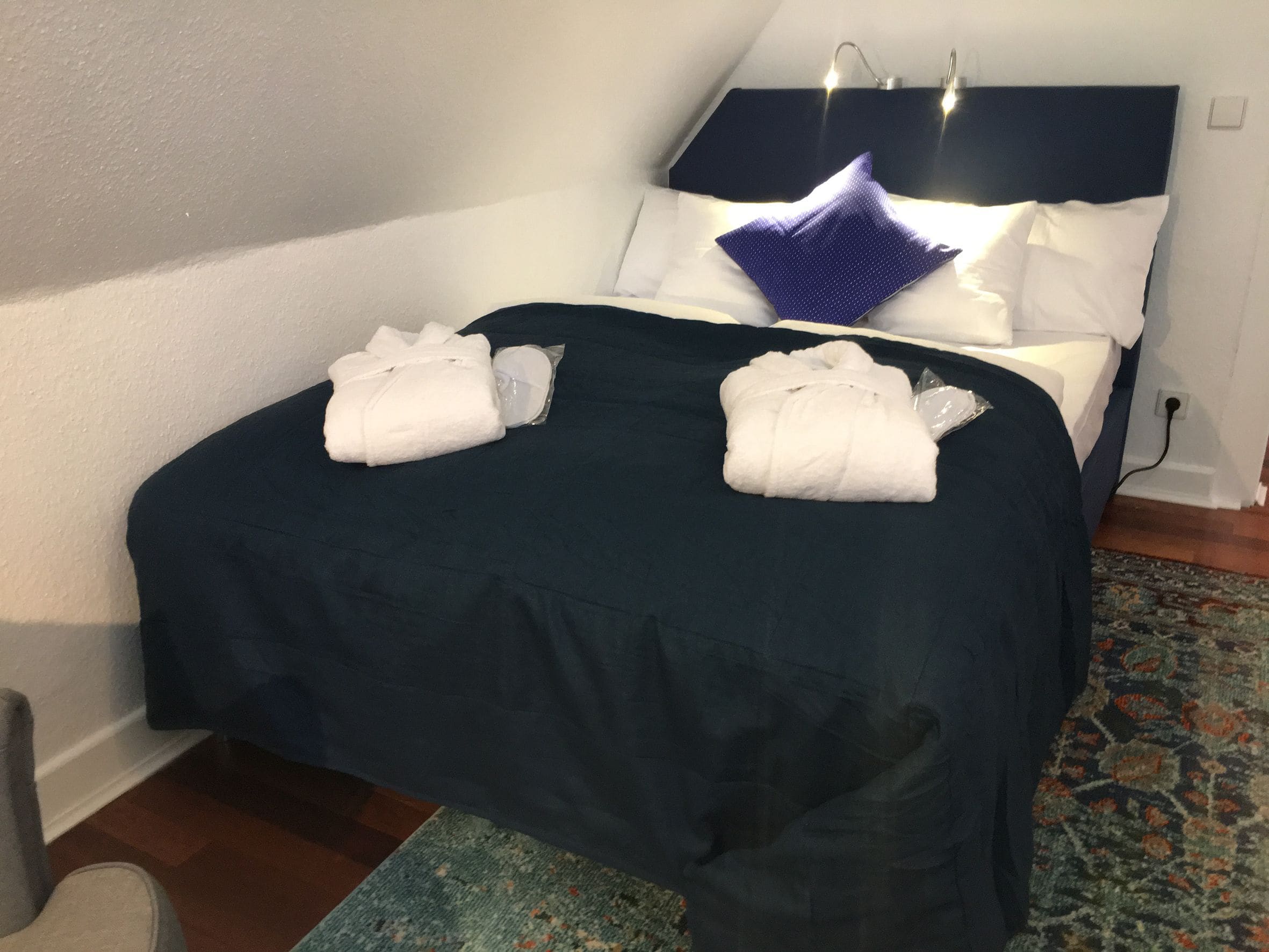 Una cama bien hecha con una colcha azul oscuro, dos toallas blancas y una única almohada azul.
