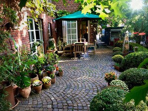 Une cour pavée de pierres, agrémentée de plantes en pot, avec une table en bois et des chaises placées sous un parasol vert.
