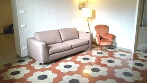 Una stanza con un divano, una poltrona, un pavimento decorato e una lampada fissata al muro.
