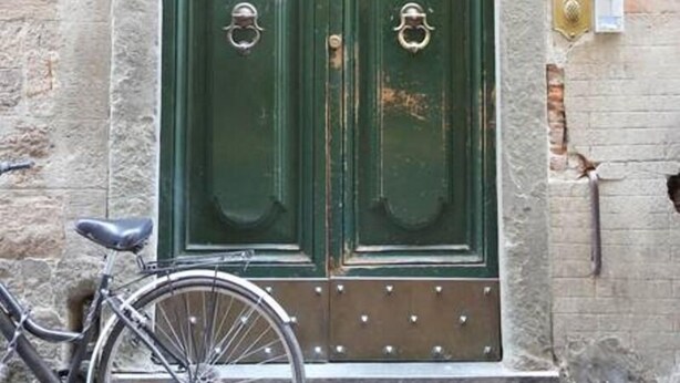 Una porta verde doppia con un arco decorativo in metallo sopra, una bicicletta parcheggiata davanti e un cartello con il numero '49' sulla parete.