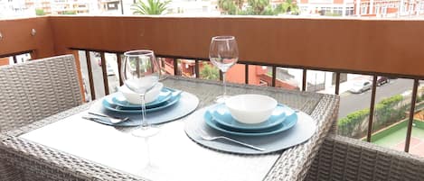A wicker table set with plates, bowls, and wine glasses on a balcony overlooking a cityscape with buildings and palm trees.
