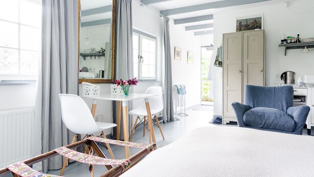 Una habitación con cama, escritorio y sillas, un espejo grande y ventana con cortinas.