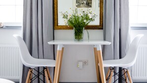 Una habitación con una mesa blanca, dos sillas blancas, un espejo con marco dorado y cortinas grises.