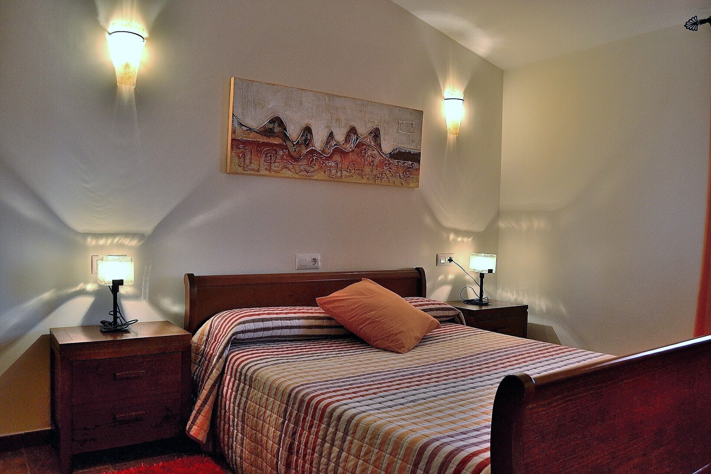 A bedroom with a bed, two bedside tables, a nightstand, a wall-mounted painting, and two wall-mounted lamps.