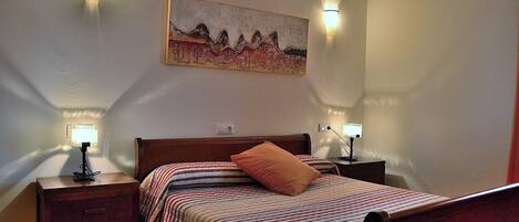 A bedroom with a bed, two bedside tables, a nightstand, a wall-mounted painting, and two wall-mounted lamps.