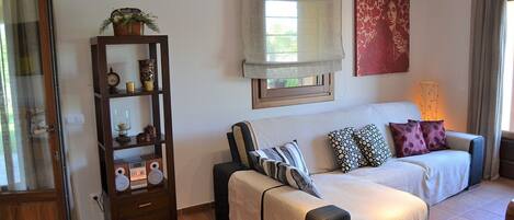 A cozy living room with a sofa set, a wooden side table with decorative items, and a window with a view.