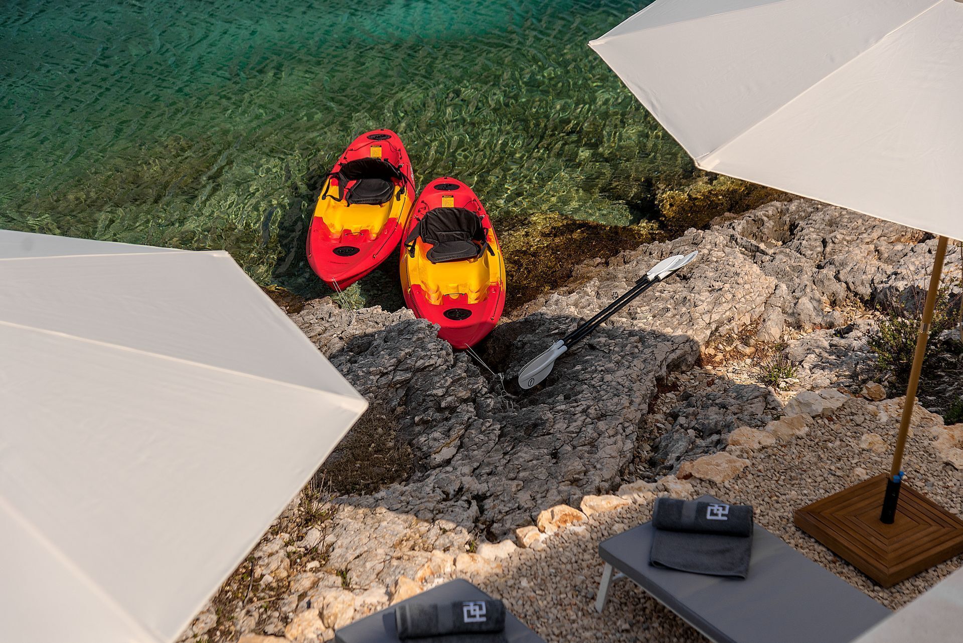 Deux kayaks sur une plage rocheuse, avec une eau claire, une pagaie et des parasols.