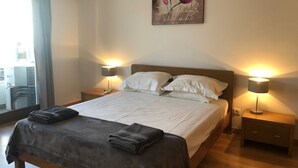 Una cama bien tendida con sábanas blancas y una manta gris oscuro, flanqueada por dos lámparas de noche sobre mesitas de madera.
