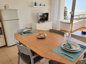 Uma sala de jantar com mesa de madeira, cadeiras, um recipiente com lanches e uma TV sobre um móvel.