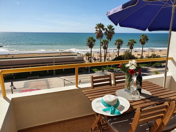 Un balcón con vistas a la playa, mobiliario de madera, una mesa con una botella y flores, y un paraguas azul.