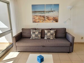 Un sofá gris con cojines estampados, una mesa blanca con un cubo azul y una pintura de una escena de playa en la pared.