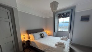 A neatly made bed with white linens and a decorative pillow, a bedside lamp, and a window with a view of the sea.