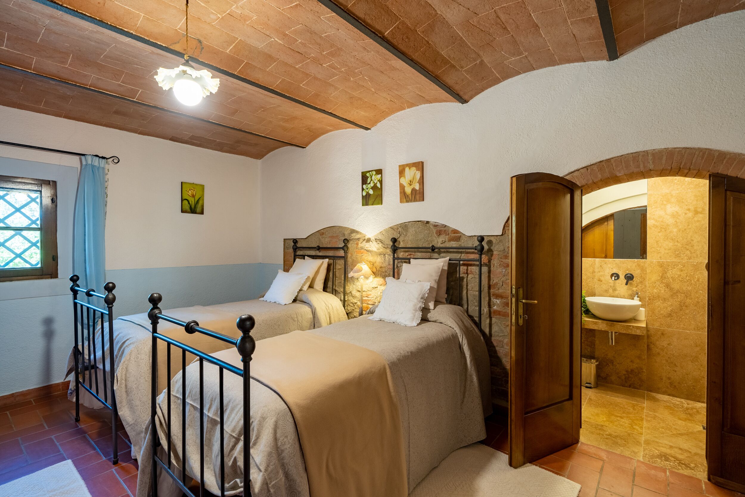 Une chambre avec deux lits, une tête de lit en pierre, une salle de bain avec un lavabo et une fenêtre avec des rideaux bleus.