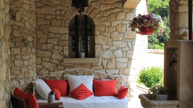 A stone-walled room with a white sofa, red cushions, a wooden chair, a hanging light fixture, and a stone fireplace with a flower pot.