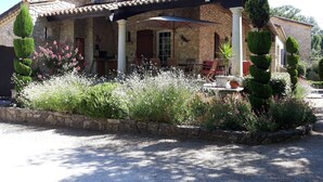 A stone building with a tiled roof, a covered patio with chairs, and a garden with various plants and flowers.
