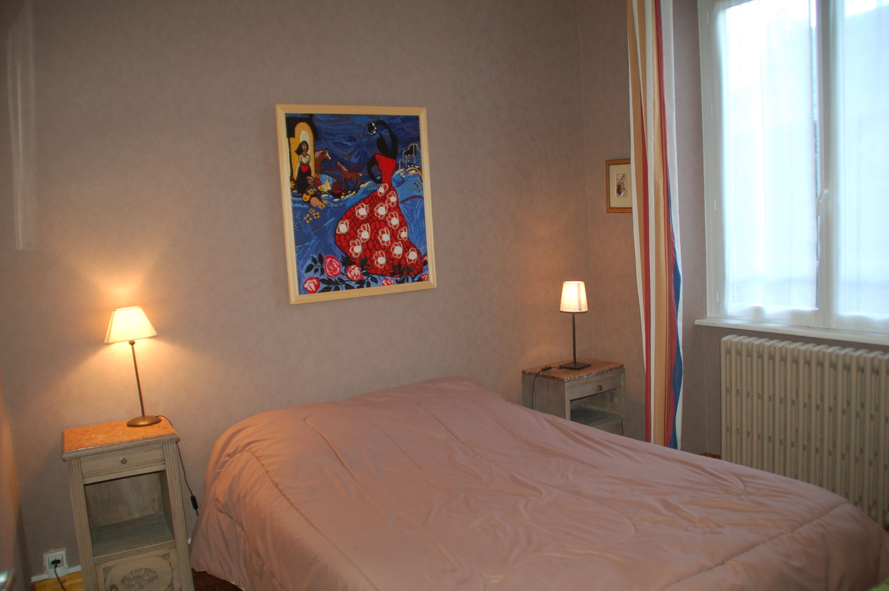 Un dormitorio con cama, dos mesitas de noche con lámparas, un cuadro en la pared y una ventana con cortinas.