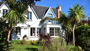 Una casa de dos pisos con fachada azul y blanca, un balcón visible y un jardín bien cuidado con plantas variadas y césped.