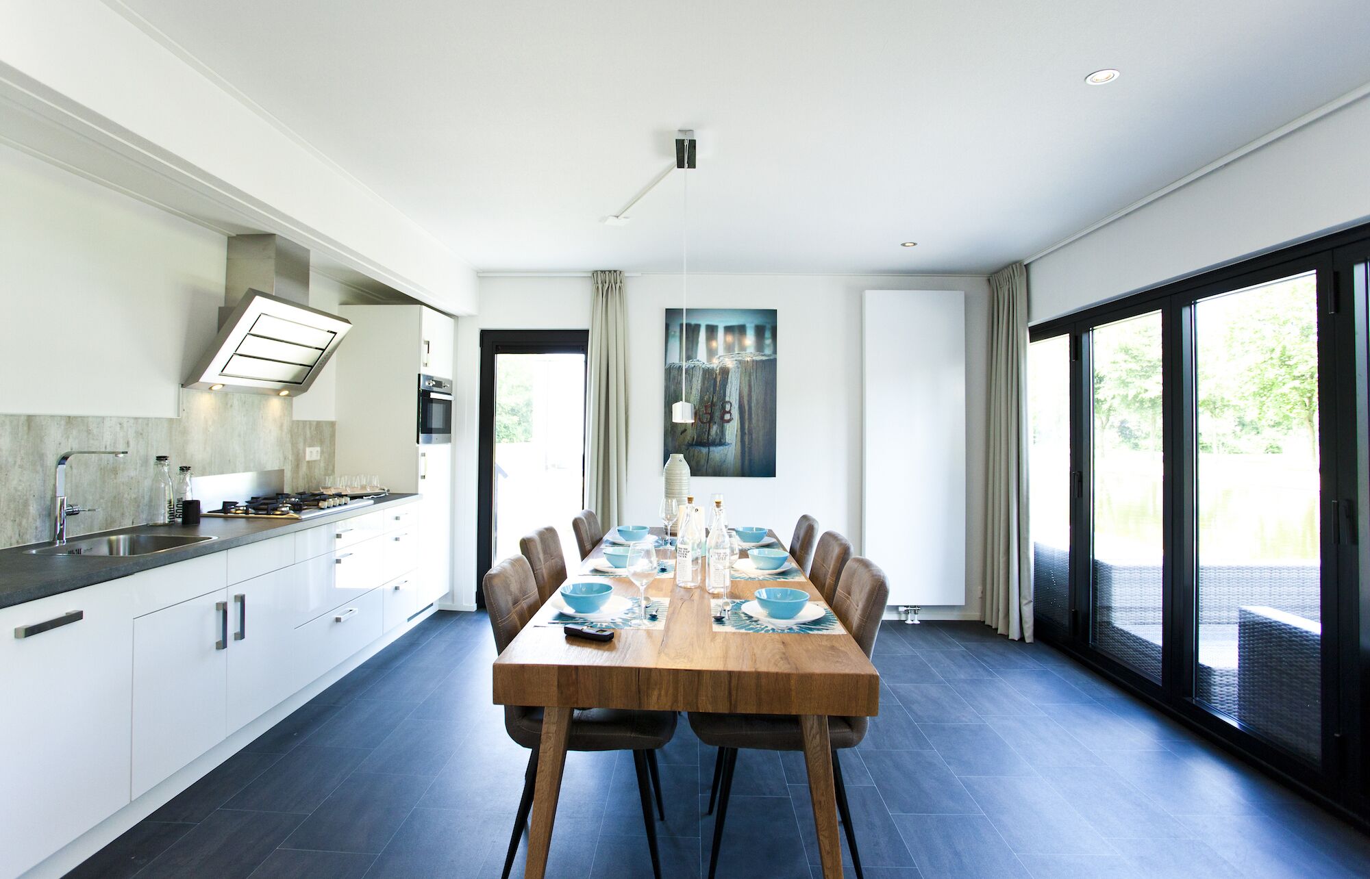 Een moderne keuken met een eethoek, met een grote tafel gedekt voor zes personen, een inbouwoven en uitzicht door de glazen deur.