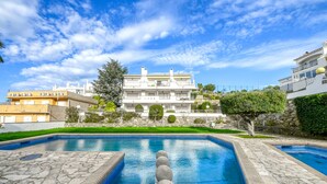 A swimming pool with stone tiles and a stone balustrade, surrounded by a well-maintained lawn and a large building with balconies. - JARDINS III 412 Roses (Rosas / Roses)