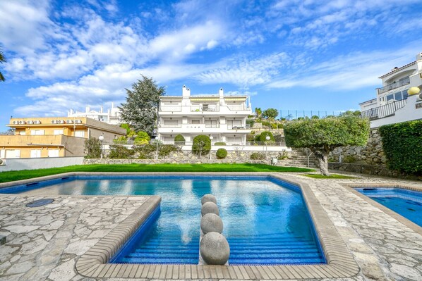 A swimming pool with stone tiles and a stone balustrade, surrounded by a well-maintained lawn and a large building with balconies.