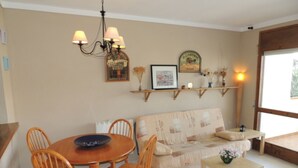 A dining area with a round table, chairs, a sofa, a small table with a vase, and wall-mounted shelves with framed items. - JARDINS III 412 Roses (Rosas / Roses)