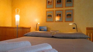 Ein ordentlich bezogenes Bett mit Kissen und aufgerollten Handtüchern, eine Nachttischlampe und gerahmte Bilder an der Wand.