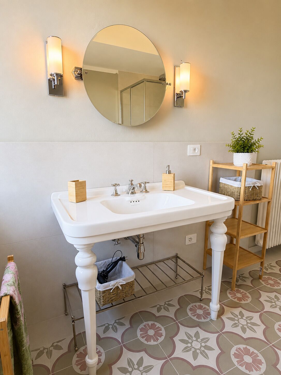 A bathroom with a white sink, two wall-mounted lights, a round mirror, a wooden shelf with baskets, and a patterned floor.