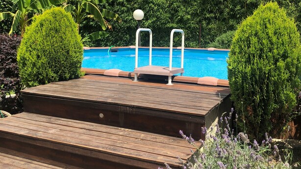 Ett trädäck som leder fram till en swimmingpool, omgiven av frodig grönska och en klarblå himmel.